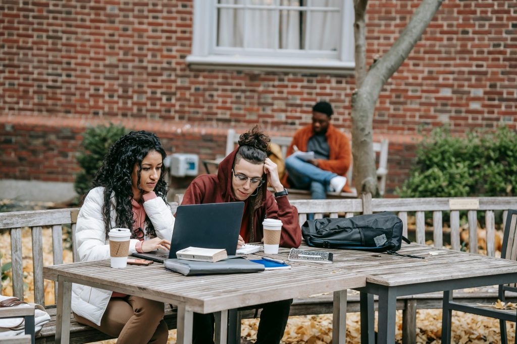 Concentrated young diverse students in casual clothes using laptop and drinking takeaway coffee while doing online project sitting at wooden table in campus park on autumn day