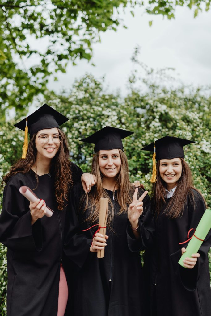 Three young women celebrating graduation with diplomas outdoors in nature.